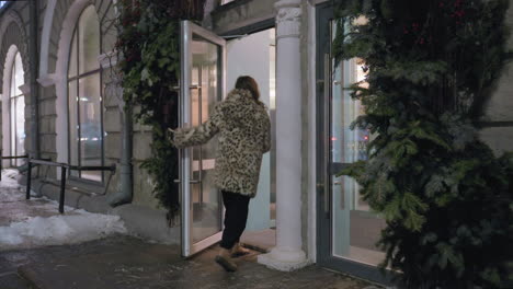 stylish woman in spotted winter jacket approaches glass door entrance surrounded by evergreen decorations on snowy evening. city lights and icy sidewalk create cozy cold weather atmosphere