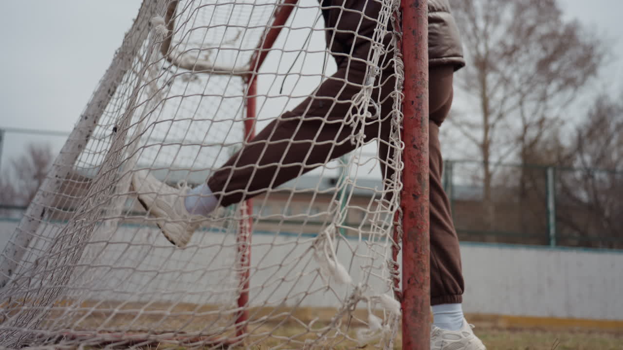 giovane giocatore appoggiato alla rete, ritratto informale di street soccer con palo arrugginito; felpa con cappuccio e scarpe da ginnastica, campo urbano nuvoloso, posa spontanea che suggerisce riscaldamento pre-partita e pausa riflessiva, editoriale urbano