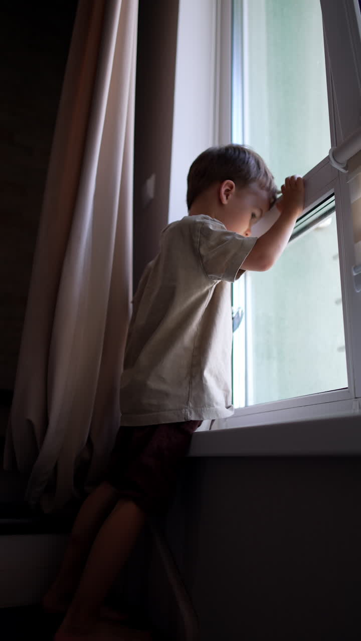 Caucasian kid wearing t-shirt and shorts stands at the window. Toddler boy leaned to the glass looking outside. Low angle view. Vertical video.