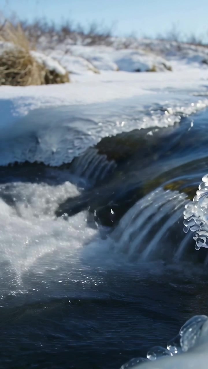 Video verticale: Acqua che risale la corrente su un piccolo salto, una cascata che forma schiuma in un ruscello con ghiaccio.