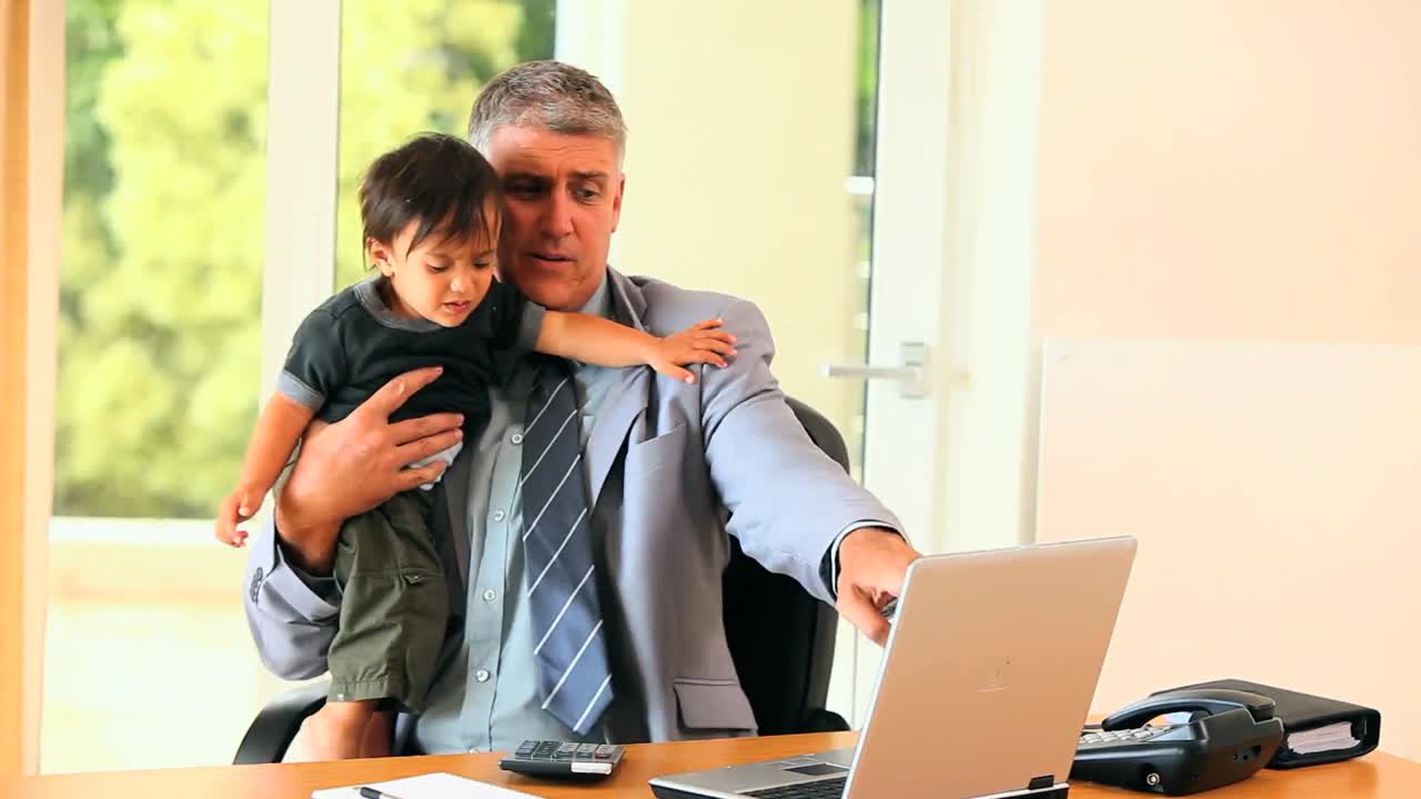 Empresario trabajando en el escritorio mientras sostiene a su hijo en brazos