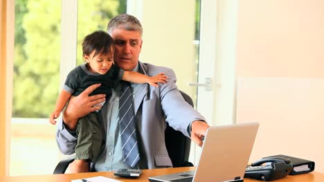 Businessman-working-at-the-desk-while-holding-his-son-in-his-arms