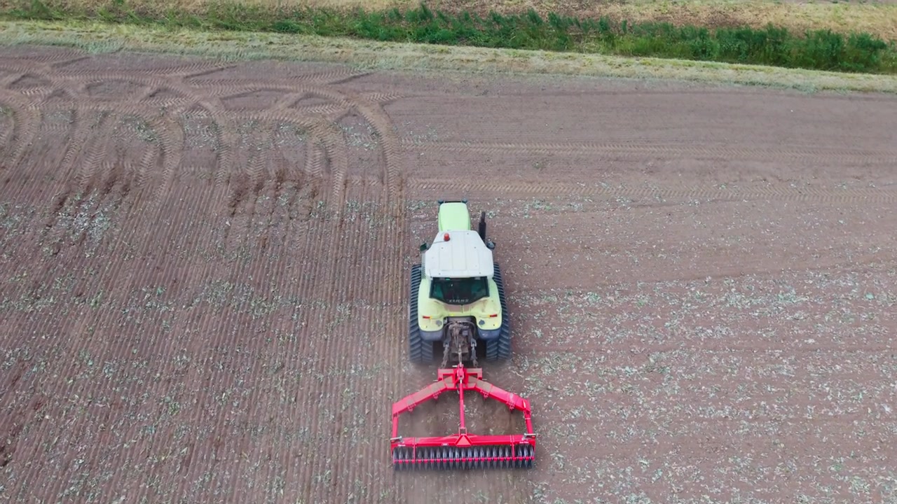 Vista aerea di un trattore che ara un campo al tramonto. Il mezzo è al lavoro su un terreno agricolo, lasciando scie di polvere e disegni nel suolo arato, per la preparazione del terreno e la coltivazione delle colture in una fattoria nel Regno Unito.