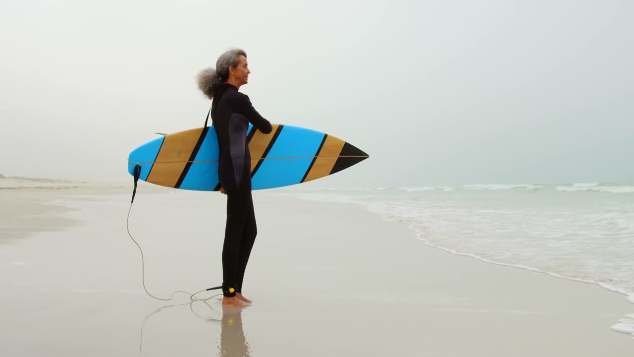 vista lateral de una surfista femenina afroamericana senior activa con una tabla de surf de pie en la playa 4k