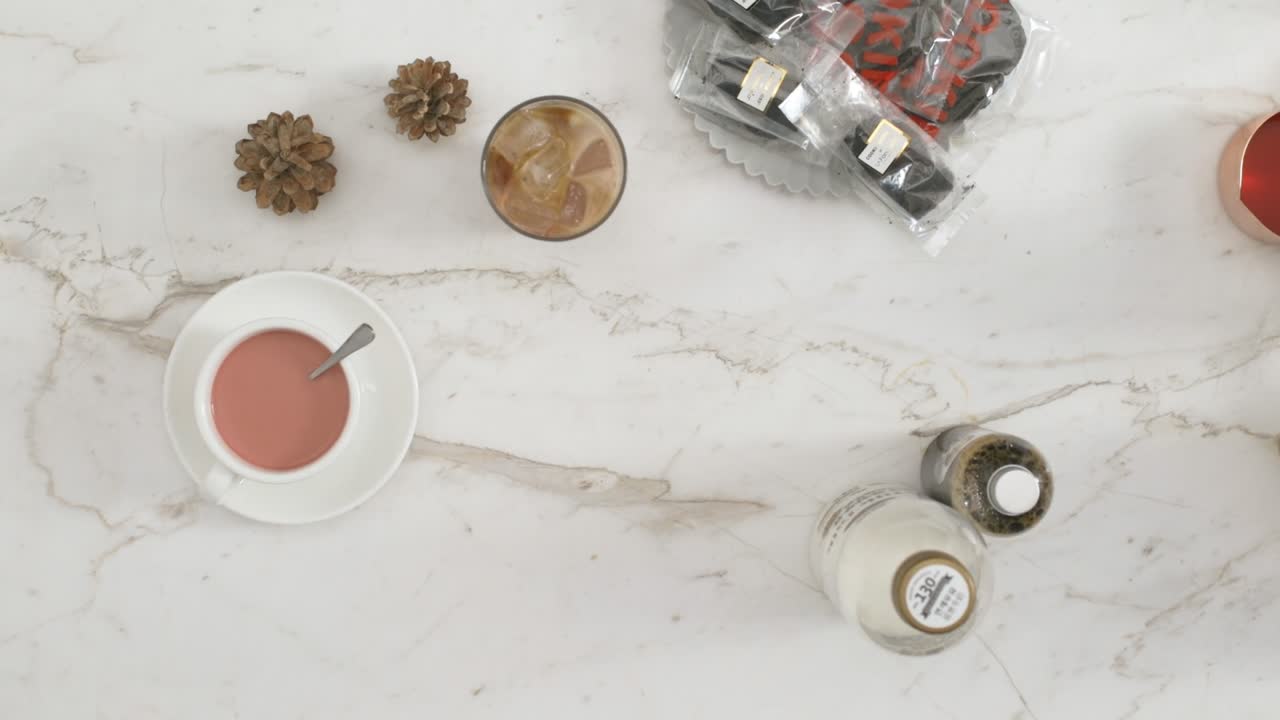 Top-down view of hands holding a cup of red velvet latte on a marble table with iced coffee, pinecones, bottled ingredients, and packaged treats.