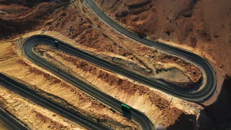 Green-bus-drives-down-a-black-tarmac-road-as-an-oncoming-car-drives-up-the-hairpin-bend-of-the-brown-mountainous-landscape-of-Mitzpe-Ramon