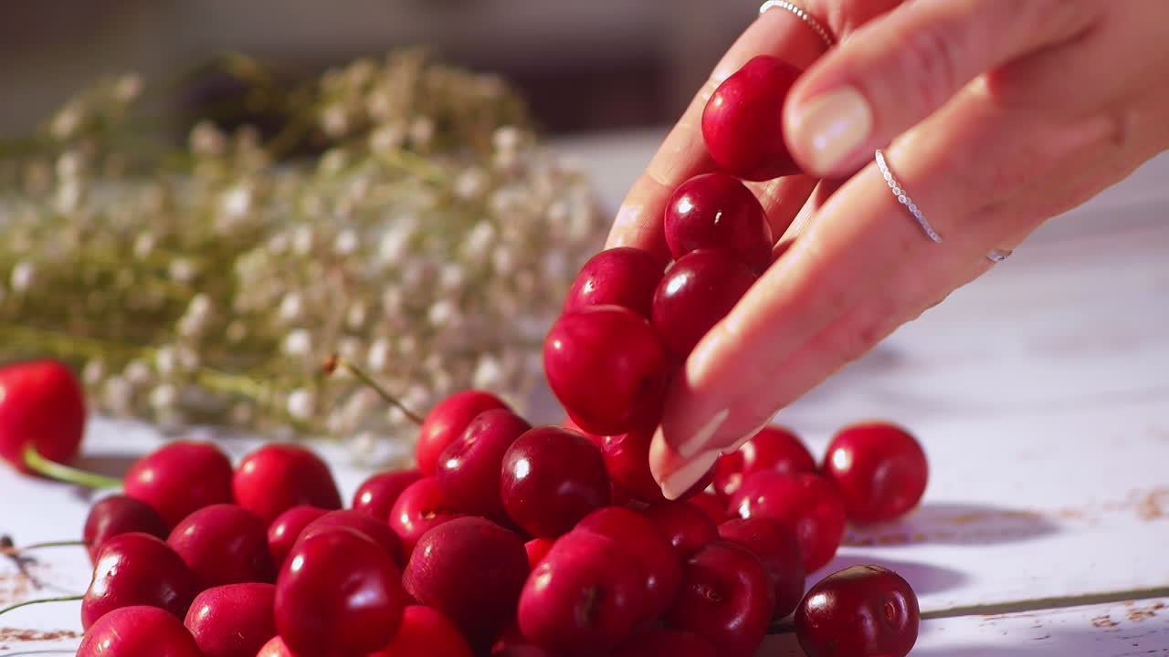 Ripe red cherries cascade in slow motion from a polished woman hand onto a white rustic table, with soft floral decor in the background adding a peaceful, summery atmosphere.