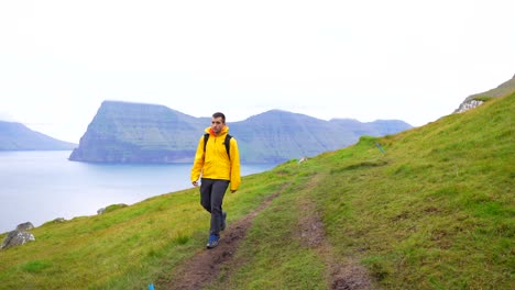 Hombre-Caminando-Hacia-La-Cámara-En-Kalsoy-Con-La-Isla-Kunoy-En-El-Fondo,-Islas-Feroe