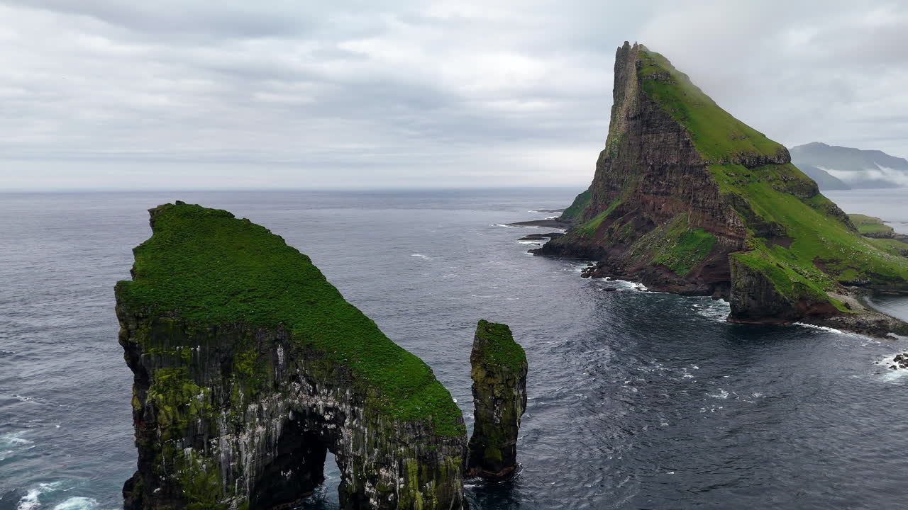 Cinematic aerial view of Drangarnir sea stacks rising dramatically from the Atlantic Ocean near Vágar, Faroe Islands, showcasing rugged cliffs, lush green slopes, and misty Nordic seascape