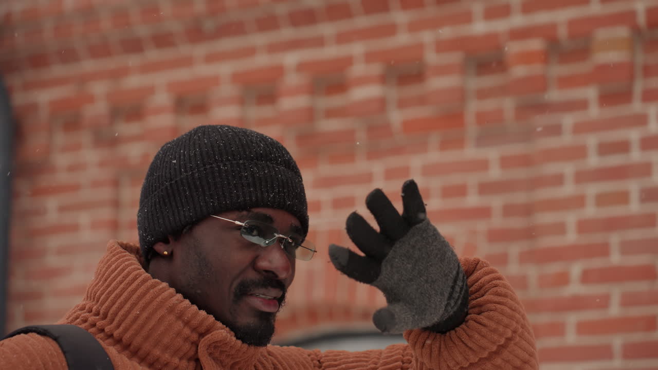 Sort mand i orange sweater vinker med løftet hånd for at skærme for genskin, smilende mod en murstensvæg. Oprigtigt urbant portræt viser hue, briller, handsker, rygsæk og en imødekommende pendlerstemning.
