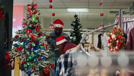 african american employees wearing santa hat ornating christmas tree in clothing store before festive promotional event. retail assitants ornating fashion shop during winter holiday season