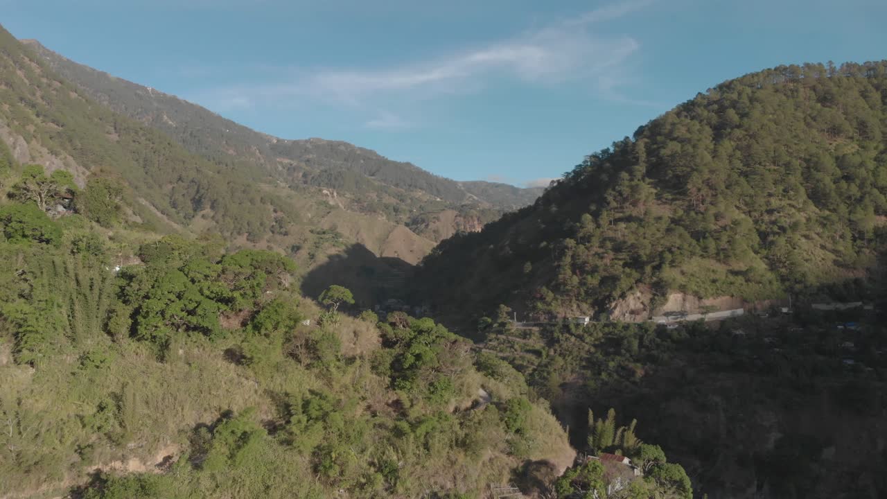 Aerial approaching and flying over a village onto a valley covered with green trees, mountains and blue sky light clouds in the background