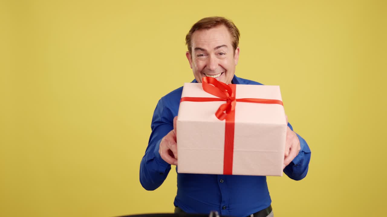Happy man offering a gift box with a smile and a wink on a yellow background