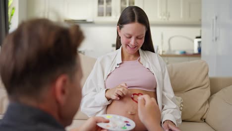 Over-the-shoulder-a-happy-man-draws-a-heart-with-bright-colors-and-a-palette-on-the-belly-of-his-pregnant-woman-at-home-during-the-weekend