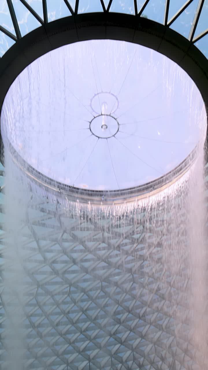 Camera tilts upward along a large indoor waterfall, revealing geometric glass dome ceiling and lush greenery in bright, natural daylight