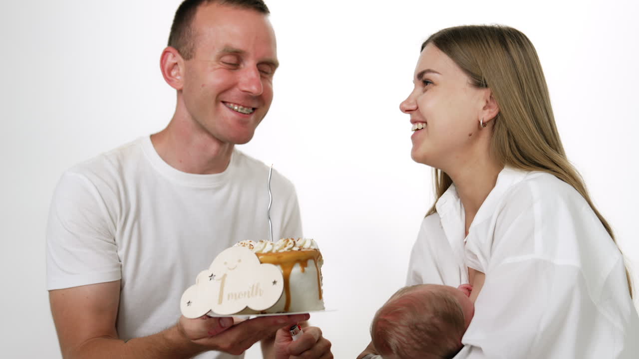 Happy family celebrating the first month of child's life with a cake. Newborn is breastfed by his mom. White backdrop.