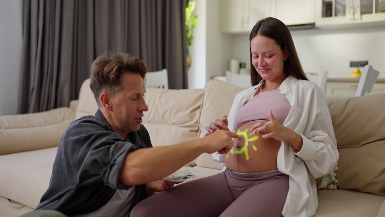 Happy brunette pregnant woman paints a sun on her belly with yellow matching paint together with her husband while expecting a baby at home