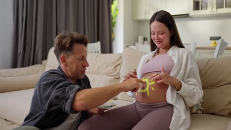 Happy-brunette-pregnant-woman-paints-a-sun-on-her-belly-with-yellow-matching-paint-together-with-her-husband-while-expecting-a-baby-at-home