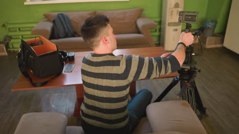 back shot of studio guy adjusting camera lens on tripod in home studio, preparing for video shoot. camera, microphone, and lighting gear are set up on table, ready for filming