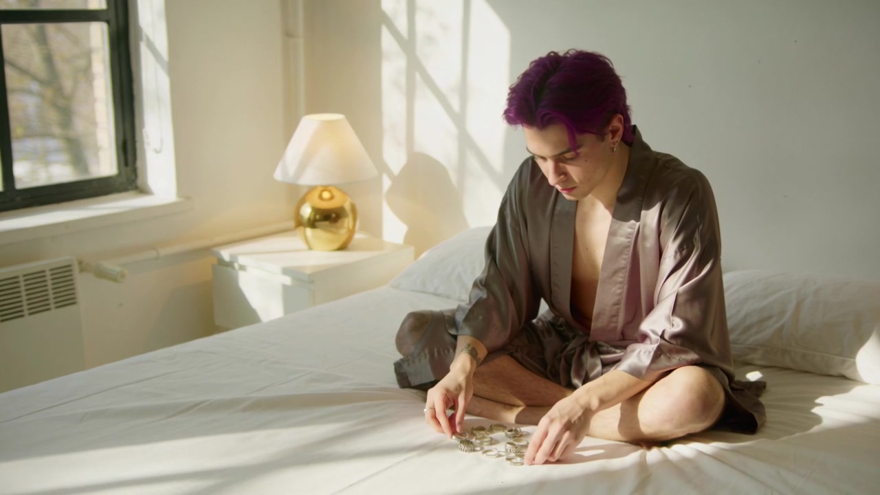 Uomo in vestaglia che tiene monete, le ordina e le conta a pile sul letto in una camera da letto illuminata dal sole, per la gestione del budget