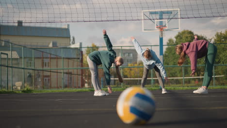 close-up of volleyball on court with athletes performing stretching exercises, hair cascading down, background features a building and greenery