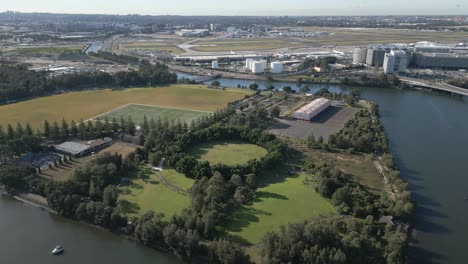 aerial zoom in flying over the cooks river, park with green trees, playground and indoor and outdoor sport stadium with sydney airport in the background