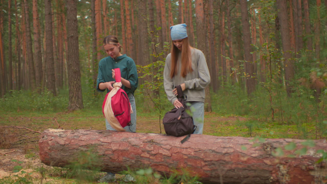 dos mujeres se acercan a un árbol caído en el bosque, sosteniendo mochilas, una con camisa verde alcanza su bolsa después de colocarla en el tronco, mientras que su compañera con un pañuelo azul abre su bolsa con atención