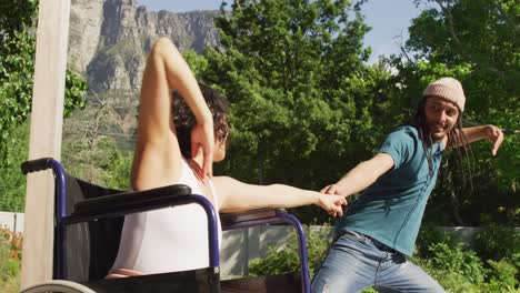 happy biracial woman in wheelchair dancing in garden with smiling male partner with dreadlocks