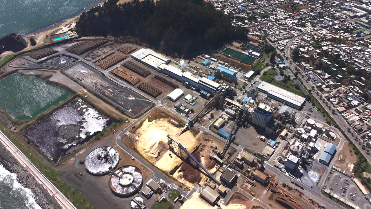 Flyover aéreo detalhado de uma planta de processamento industrial costeira no Chile, mostrando matérias-primas de madeira, tanques de armazenamento e a proximidade da instalação com a cidade residencial local