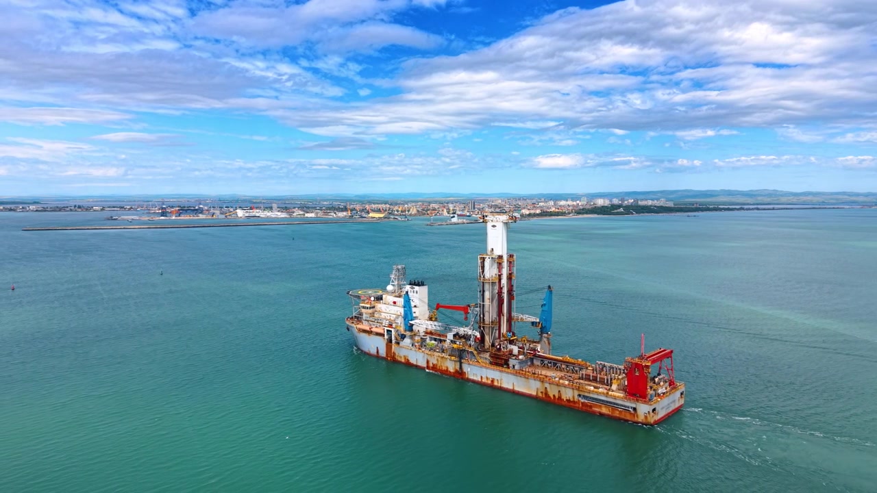 Luftfoto af et offshore boreskib, der sejler nær en kystbyhavn. Industrielt boreskib med centralt boretårn navigerer i turkist hav nær en havnekystlinje under en overskyet himmel.