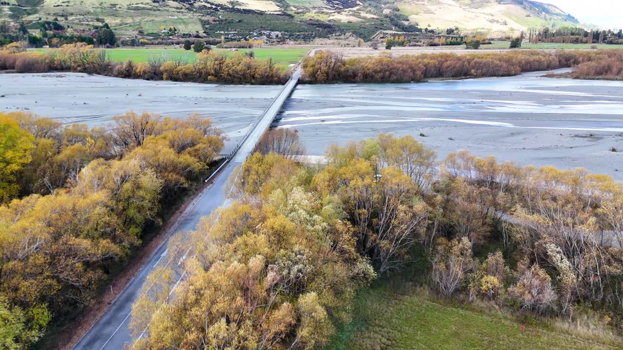 Drone captures Glenorchy Bridge amidst vibrant autumn foliage and serene river landscape under soft daylight