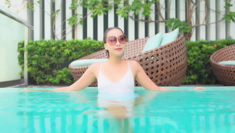 mientras se inclina a lo largo del borde de una piscina, una mujer que usa lentes de sol grandes cambia la posición de sus brazos de en el agua a lo largo del borde de la piscina