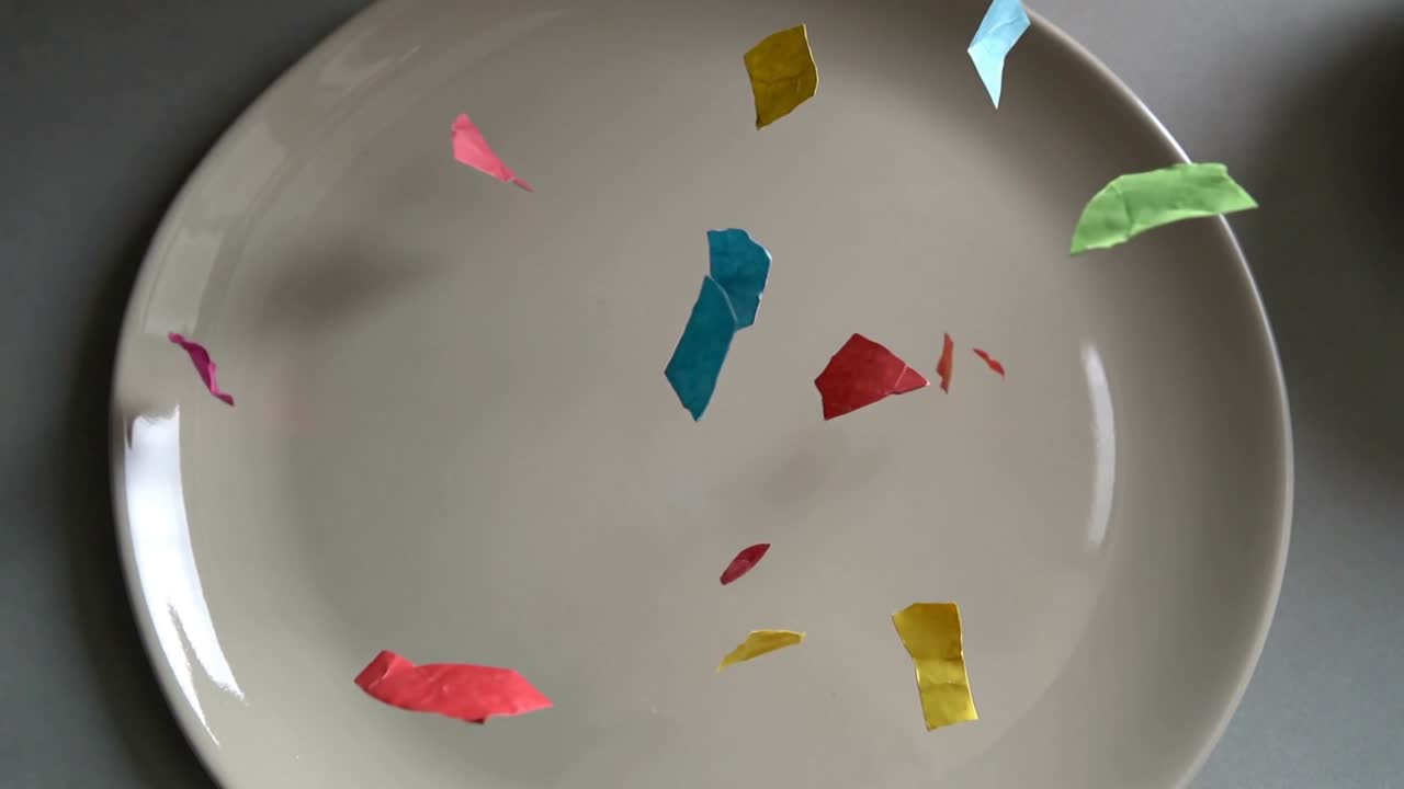 Pezzetti di carta colorata iniziano a cadere verso un piatto bianco su un tavolo grigio, posandosi, con il bordo del piatto visibile.