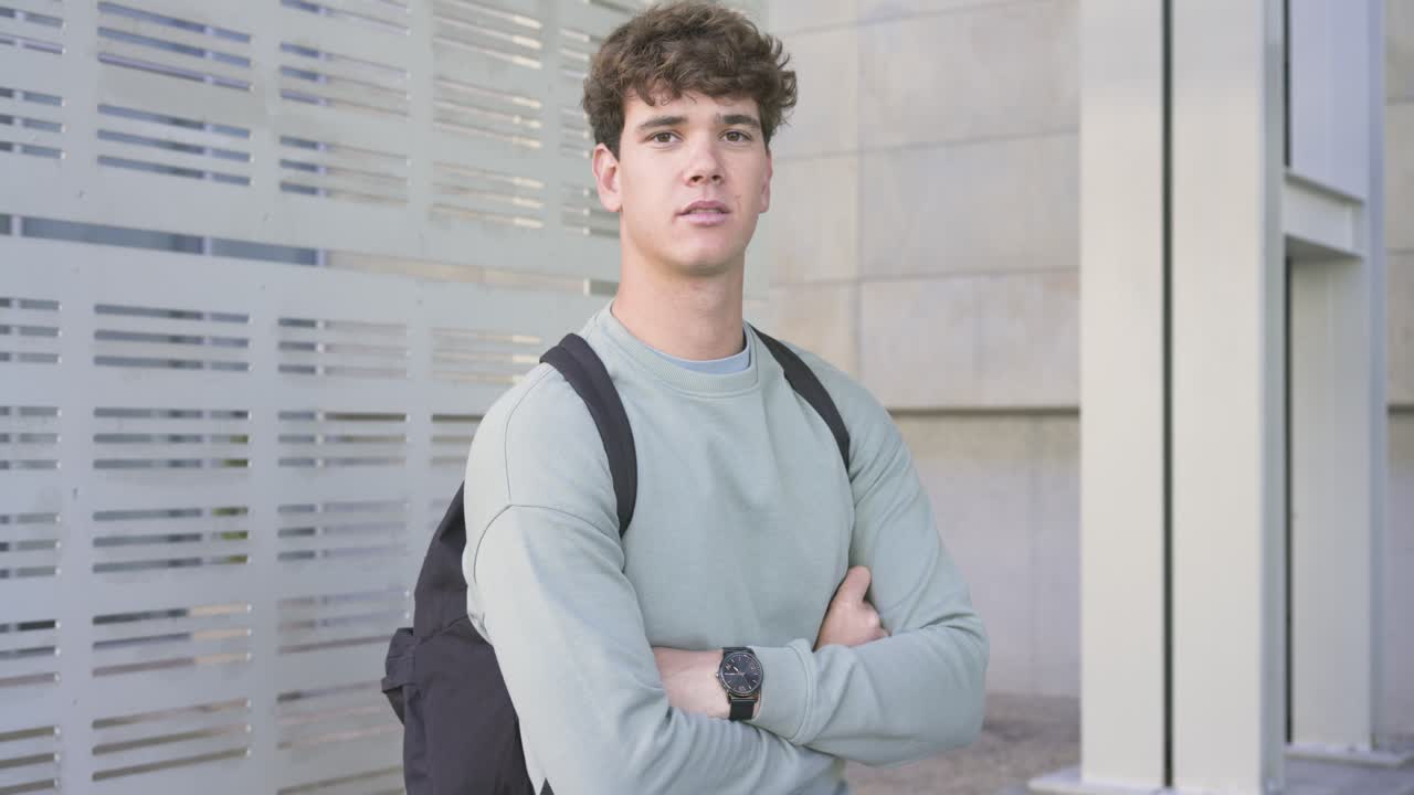 Estudante posando, se ajeitando, cruzando os braços e relaxando, enquanto a câmera se aproxima no campus, com mochila.