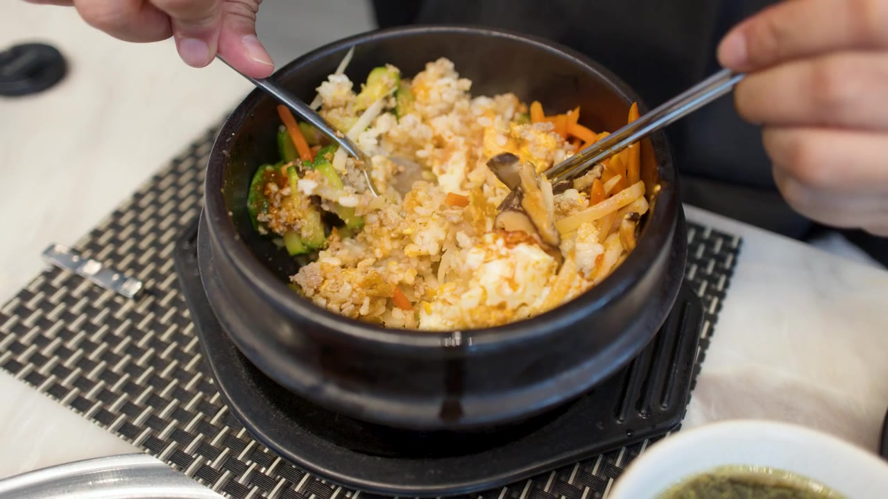 Potret jarak dekat tangan menggunakan sendok dan sumpit untuk mengaduk bibimbap dalam mangkuk batu