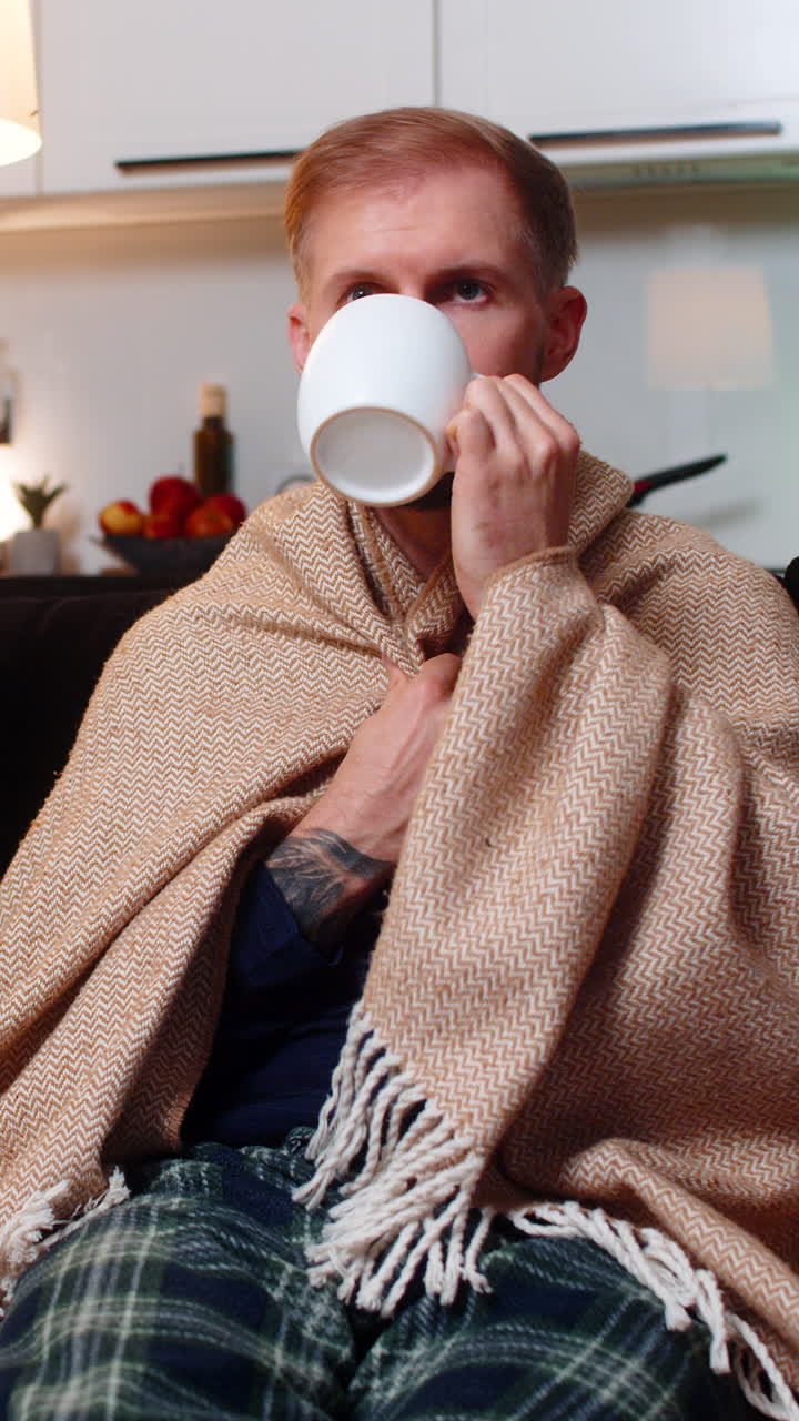Giovane uomo sul divano si scalda le mani su una tazza di tisana, ne respira l'aroma, sorseggia avvolto in una coperta e sorride
