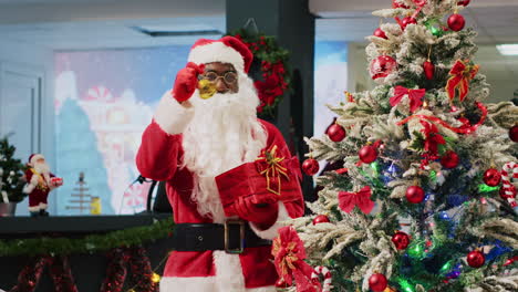 Empleado-Afroamericano-Haciéndose-Pasar-Por-Papá-Noel-Tocando-Campanas-Junto-A-Un-árbol-De-Navidad-Bellamente-Decorado,-Tratando-De-Atraer-Clientes-A-La-Tienda-De-Moda-Durante-La-Temporada-Promocional-De-Vacaciones