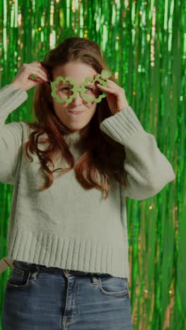 vertical video of woman celebrating st patrick's day standing in front of green tinsel curtain wearing prop shamrock shaped glasses