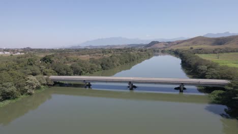 side aerial view of the bridge over the river passing by a lonely car on a sunny blue sky day with mountains in the background