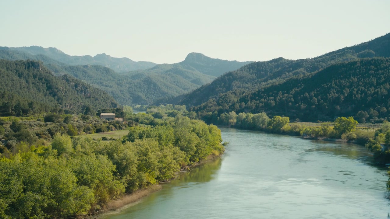 The Ebro River winds through green riverbanks and forested hills in Spain, creating a broad natural landscape with calm water and distant mountain ridges.