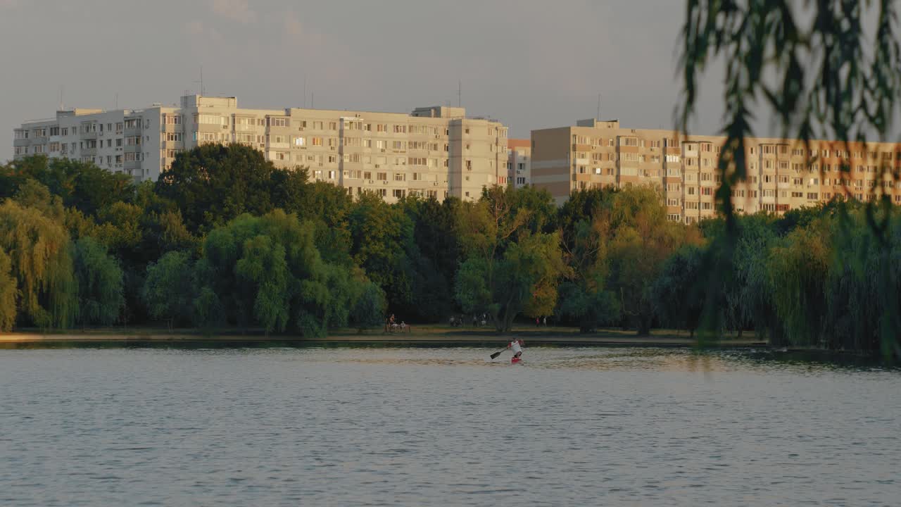 un kayak pasando por un lago con algunos viejos edificios comunistas y árboles en el fondo al atardecer