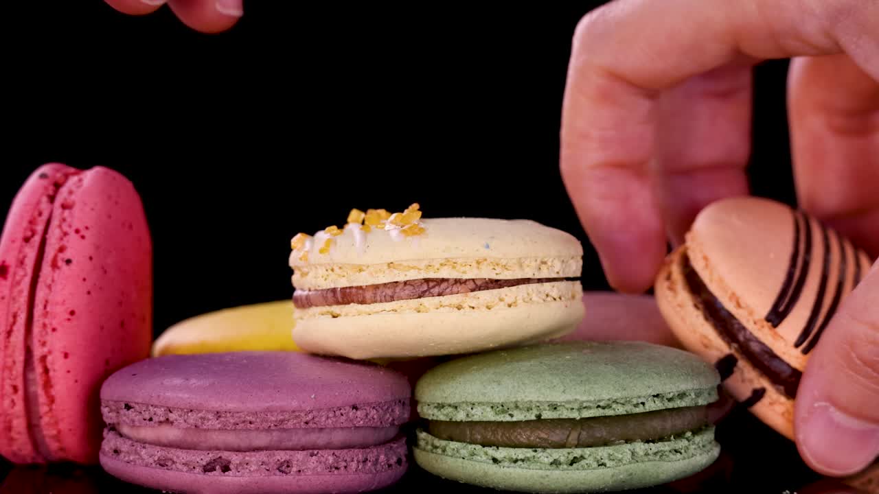 A hand places assorted pastel macarons on a reflective black surface, creating a neat stack. Bright, even lighting highlights the vibrant dessert colors and smooth textures