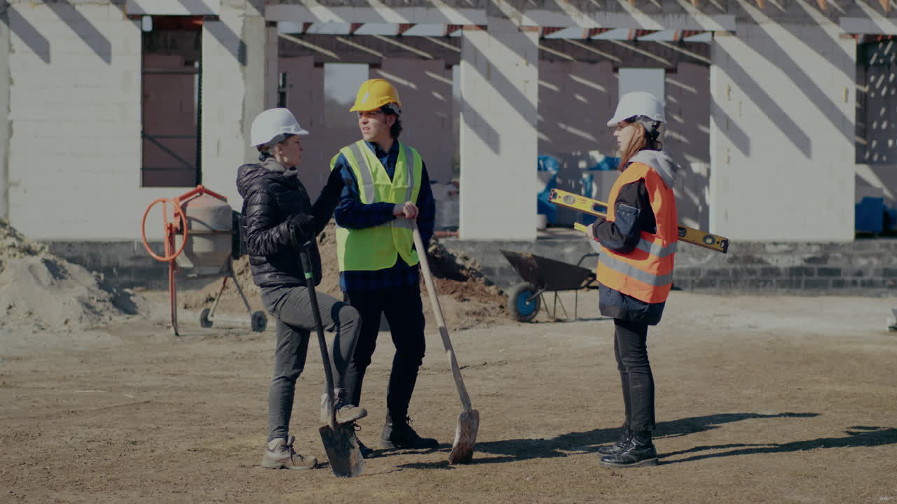 Full length of building contractor discussing with construction coworkers holding shovels standing at incomplete site