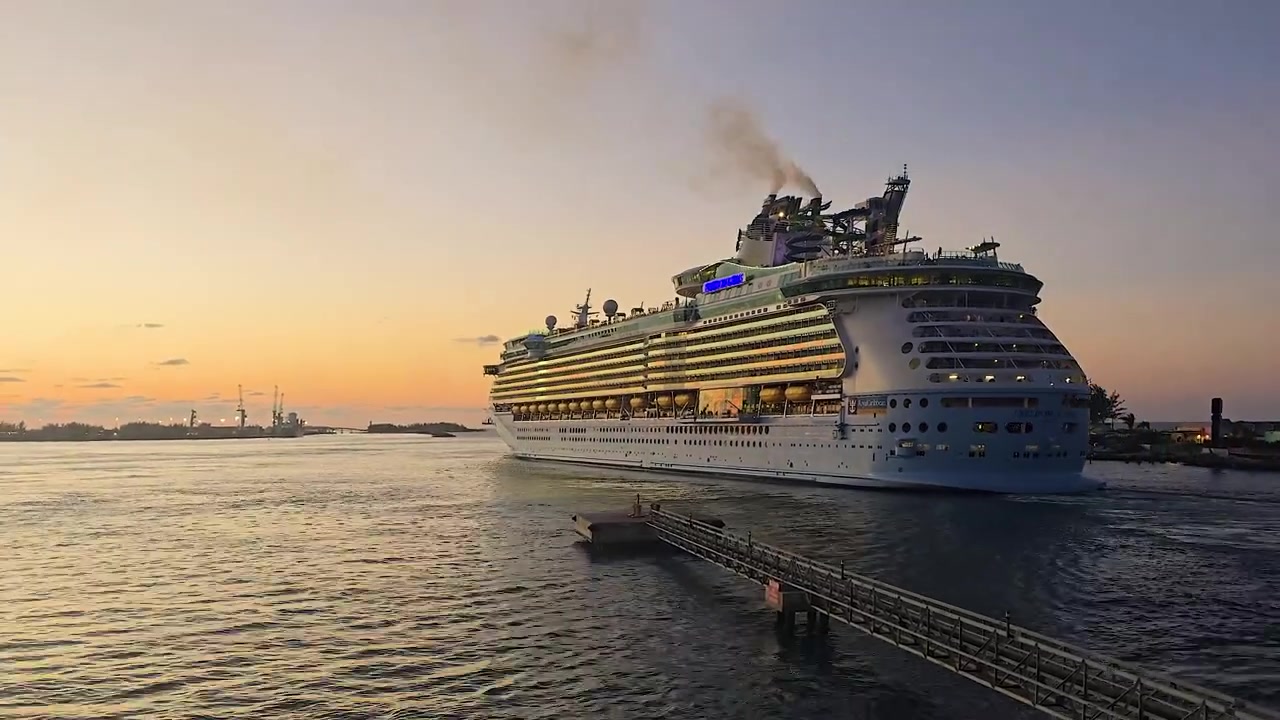 En naturskøn timelapse af et stort passagerkrydstogtskib, der afgår fra en havneterminal og sejler ud i havet under en smuk solnedgang. Fartøjet forlader havnen under en farverig aftenshimmel.