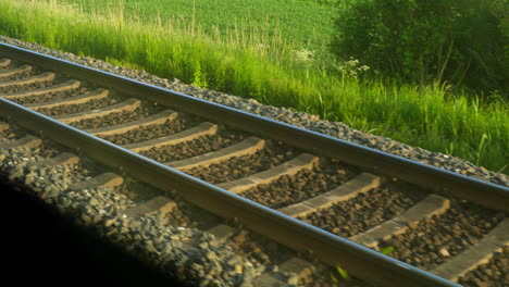 Close-Up-of-Train-Tracks-through-Train-Window-on-a-Sunny-Day,-Lush-Green-Grass-in-Background