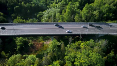 Convoy-of-vehicles-in-pursuit-over-highway-bridge-through-lush-green-forested-landscape,-fast-tracking-drone-shot-follows-from-above