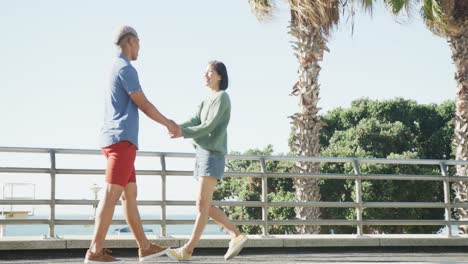 Happy-biracial-couple-walking-together-and-holding-hands-on-promenade,-in-slow-motion
