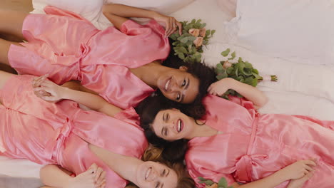 top view of group of multiethnic female friends and bride dressed in pink and white silk nightdresses holding hands with bouquets and laughing while laying on floor 2