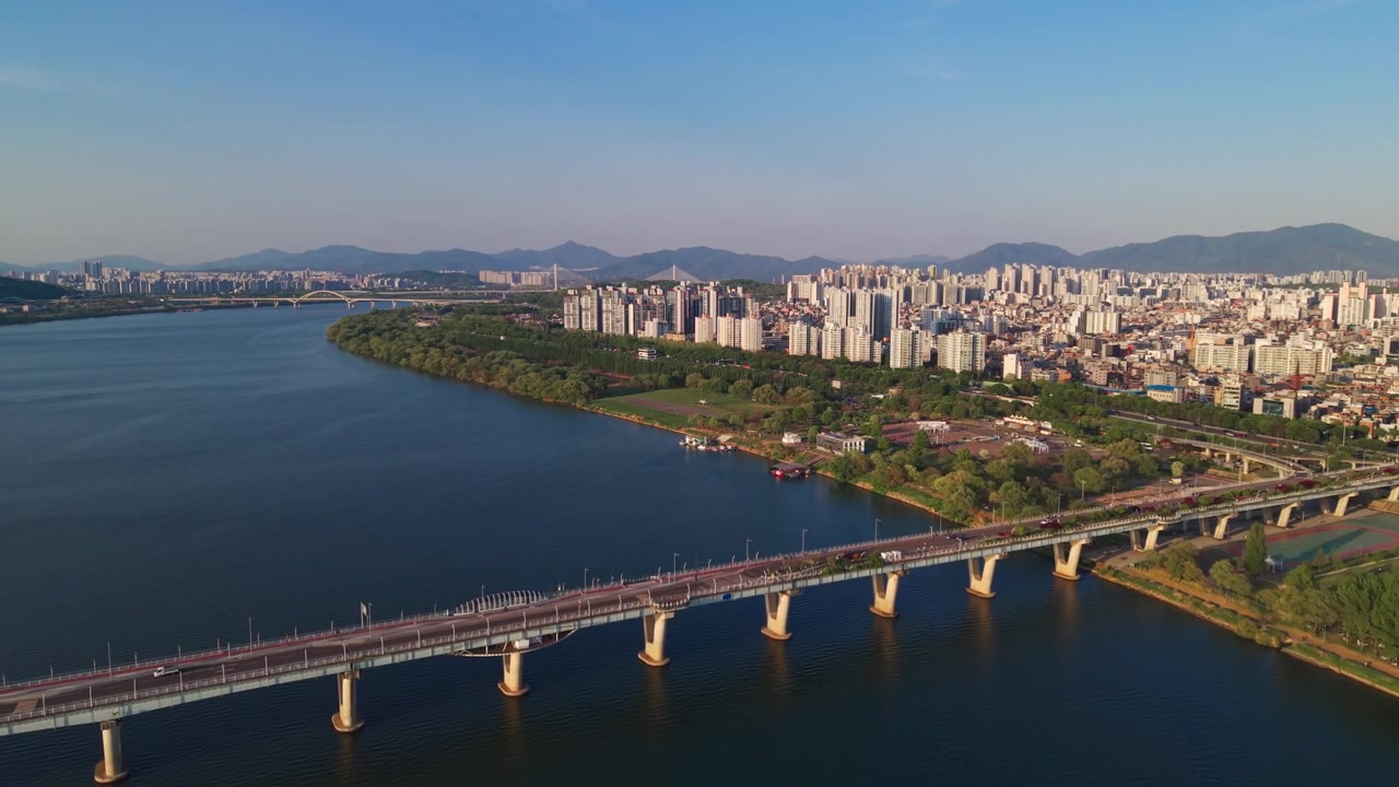 Cảnh quay từ trên cao qua cầu Gwangjin và Cheonho với giao thông chuyển động, để lộ bờ sông xanh của Công viên Gwangnaru Hangang dưới ánh nắng xuân ấm áp ở Seoul
