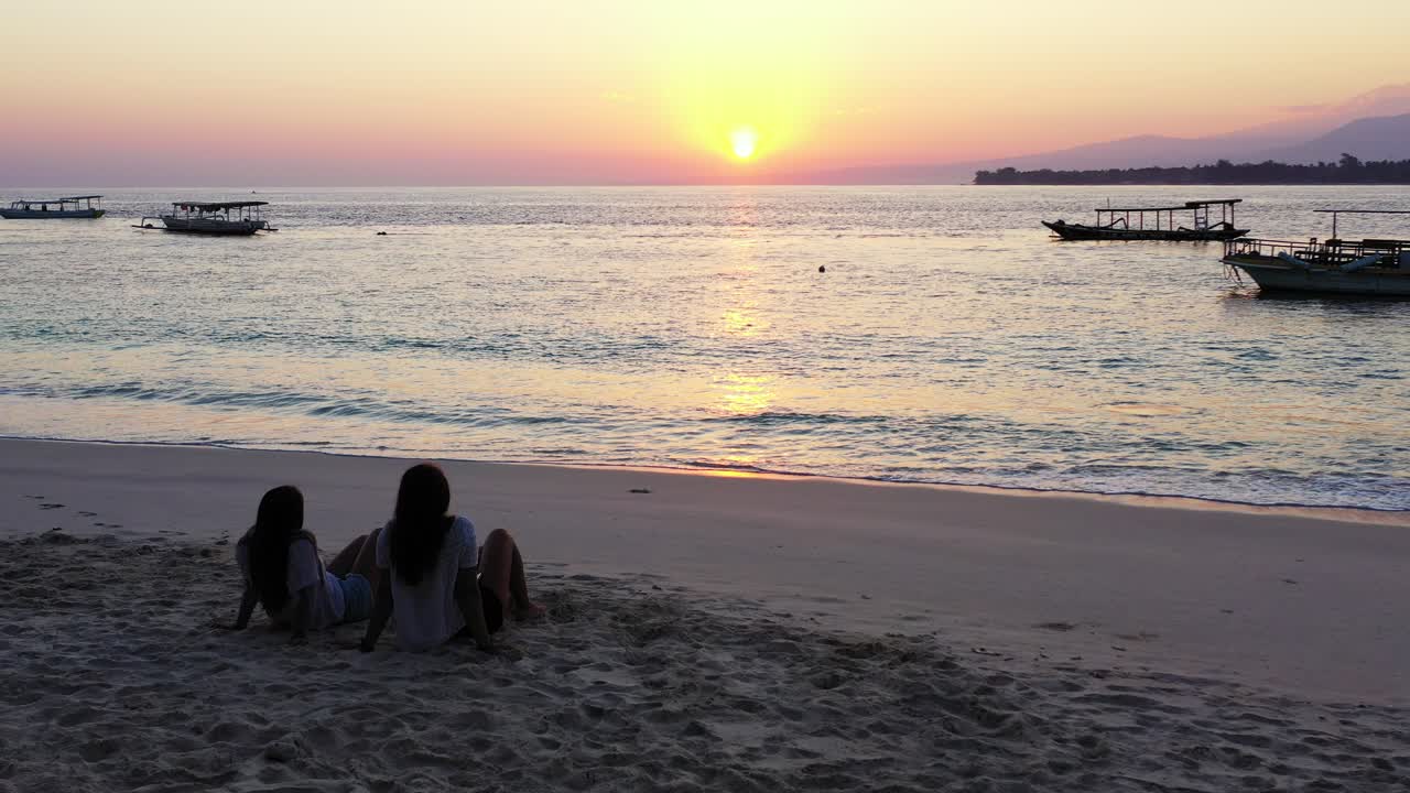 Young women sitting on sandy exotic beach, watching beautiful sunset reflecting sunlight on calm sea surface, silhouette of anchored boats